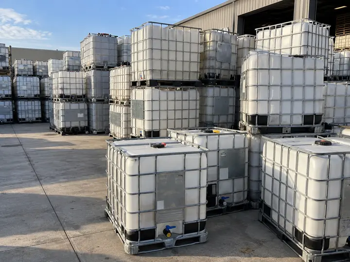 Rows of white caged IBC totes stacked two high on a concrete apron beside an open warehouse door.